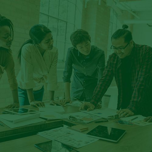 Grupo de jovens profissionais reunidos em mesa de trabalho, discutindo ideias e analisando documentos em ambiente moderno e colaborativo.