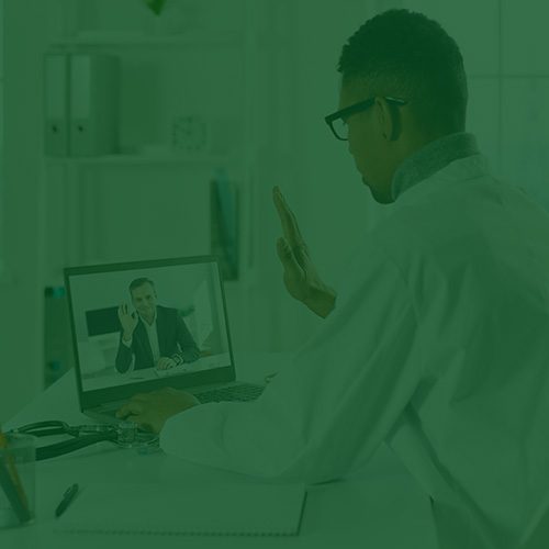 Médico com jaleco conversa por videochamada com outro homem, ambos acenando pela tela do notebook em um consultório.
