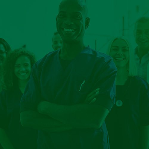 Profissionais de saúde sorrindo em grupo, com destaque para um homem de braços cruzados à frente, vestindo uniforme médico.