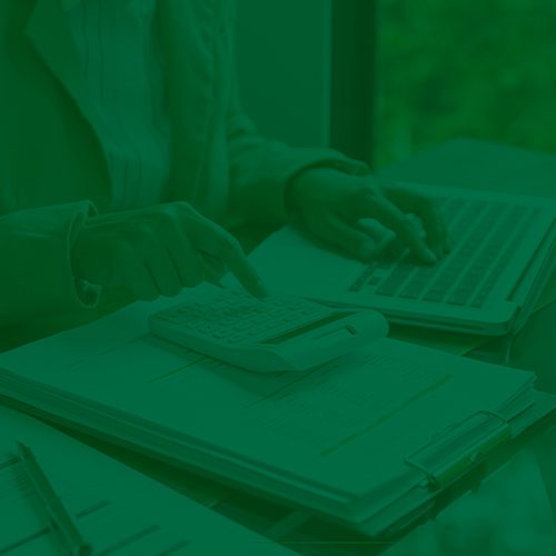 Pessoa usa calculadora sobre documentos e laptop, representando controle financeiro ou gestão de custos empresariais.