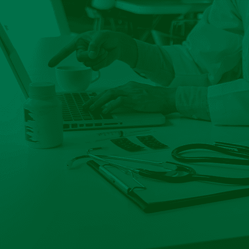 Médico usando laptop em consultório, com estetoscópio, medicamentos e receituário sobre a mesa durante atendimento.