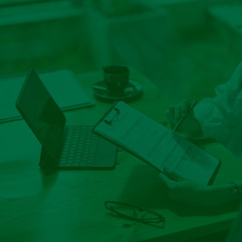 Pessoa analisando formulários em prancheta ao lado de um laptop sobre a mesa, com filtro verde sobre a imagem.