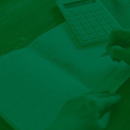 Pessoa anotando informações em planilha ao lado de uma calculadora sobre a mesa, em momento de organização financeira.